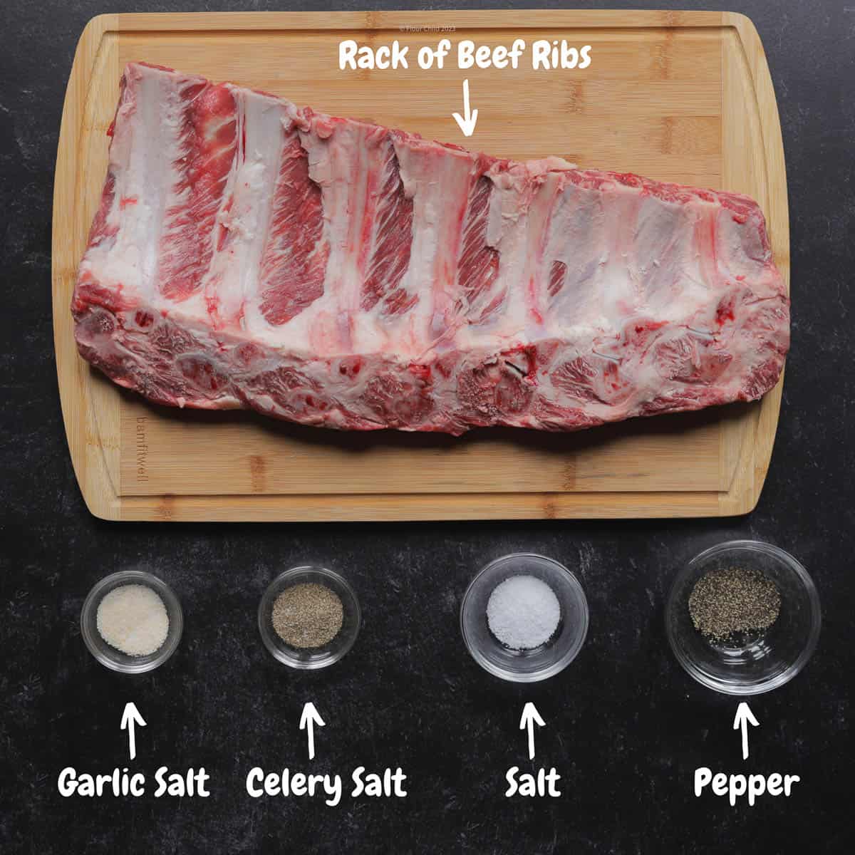 An uncooked slab of beef ribs on a wooden cutting board with the seasonings for the rub below it.