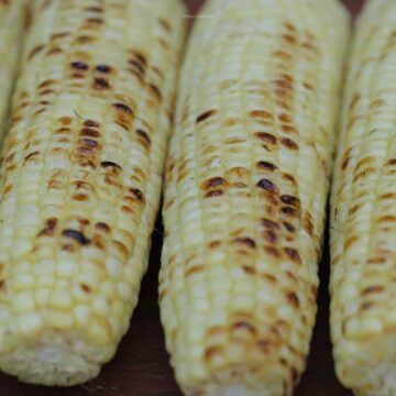 Four ears of browned, grilled corn in a row on a wooden serving tray.
