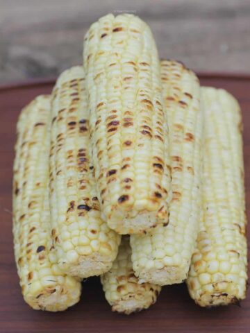 Six ears of grilled, browned corn stacked on a wooden serving tray.