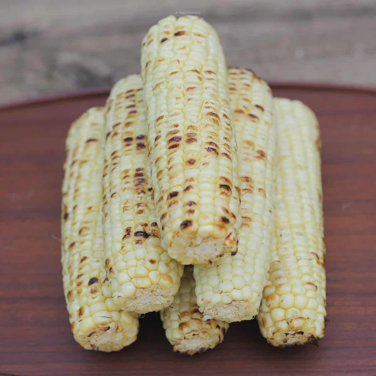 Six ears of grilled, browned corn stacked on a wooden serving tray.