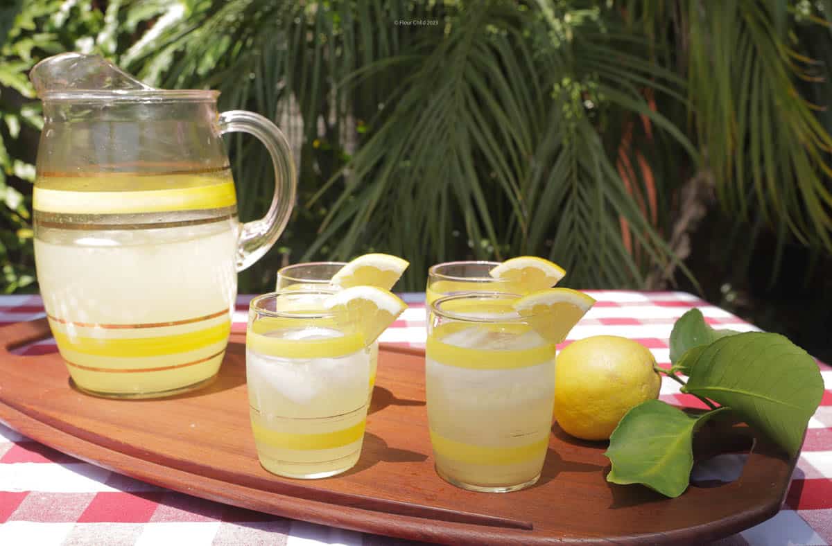 A pitcher of homemade lemonade with 4 glasses full, sitting on a wooden platter next to a lemon.