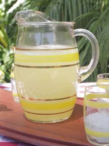 A pitcher of old fashioned lemonade ready to pour into the 4 surrounding glasses that are garnished with a lemon wedge.
