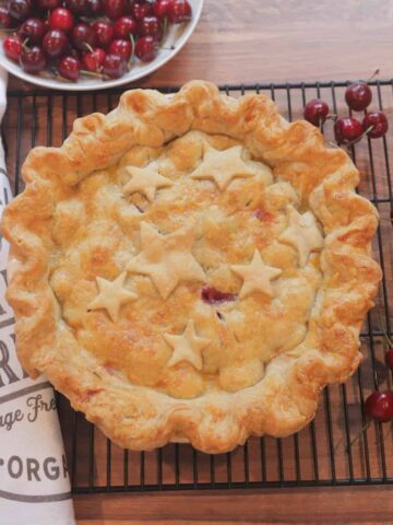 Cherry pie on a cooling rack surrounded by fresh cherries, and with a white bowl of cherries and a white kitchen towel next to it.
