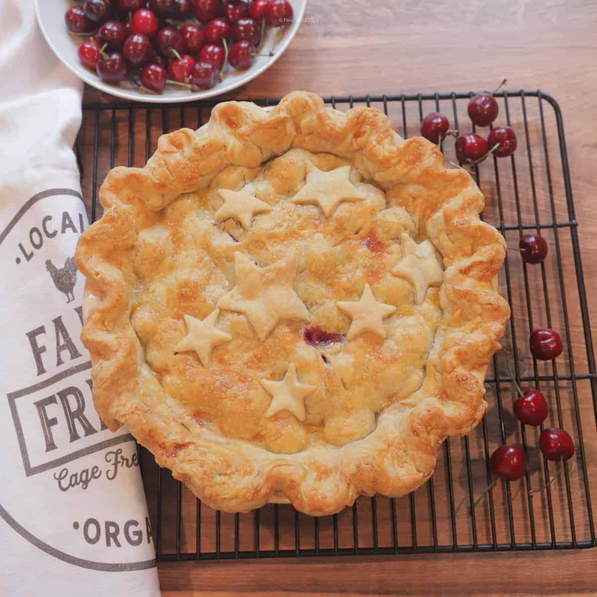 Cherry pie on a cooling rack surrounded by fresh cherries, and with a white bowl of cherries and a white kitchen towel next to it.