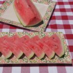 A rectangular serving plate with 7 watermelon wedges stacked, with a large square serving plate behind it and one quarter of a whole watermelon on it.
