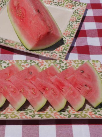 A rectangular serving plate with 7 watermelon wedges stacked, with a large square serving plate behind it and one quarter of a whole watermelon on it.