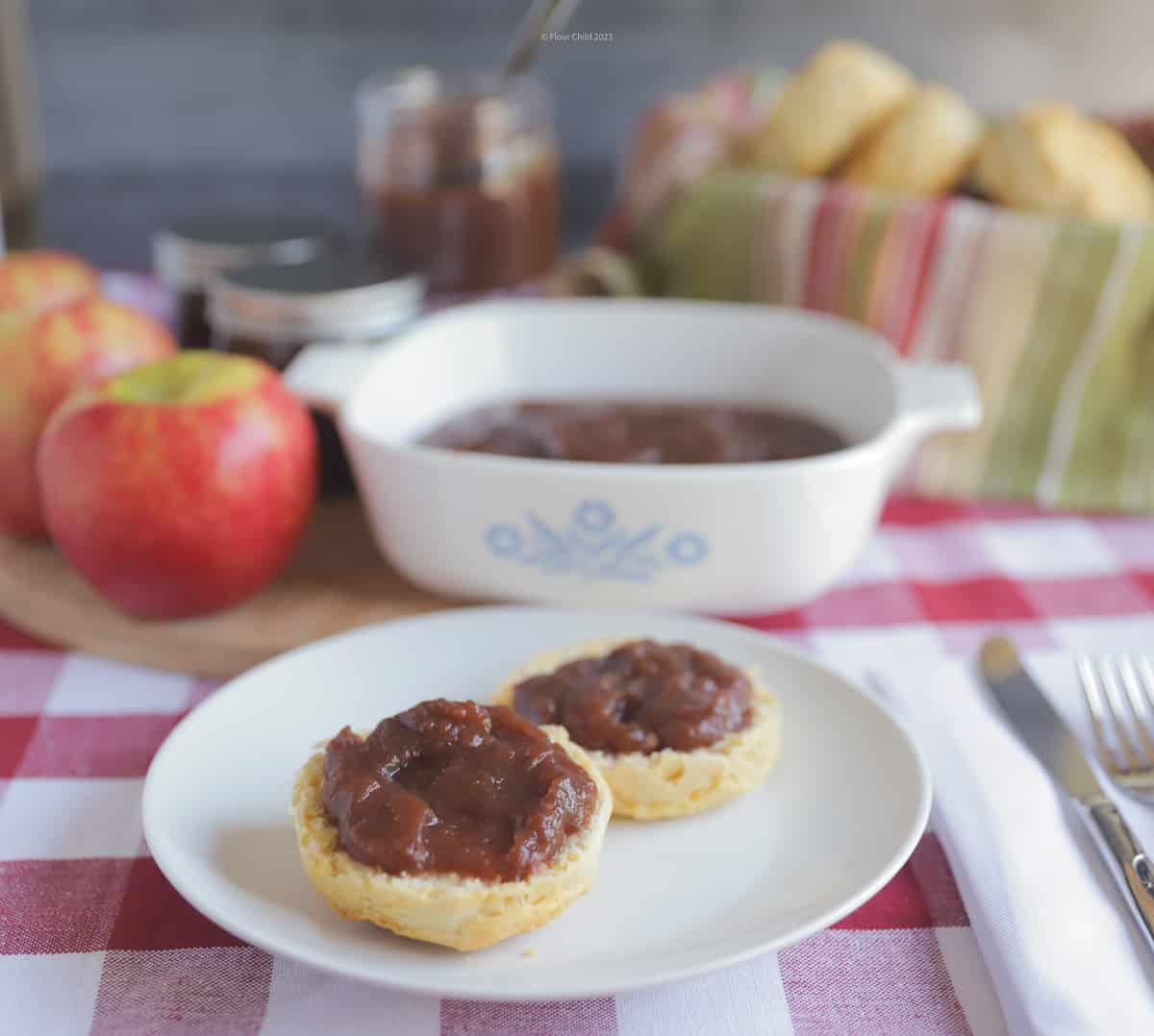 Apple butter on top of a biscuit on a white plate, with the remainder of the apple butter in a Corningware bowl behind it. 