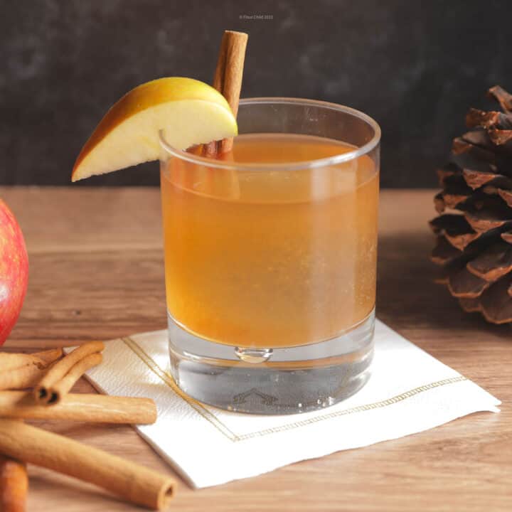 An apple butter old fashioned cocktail in a tumbler with apples and fall-related items in the background