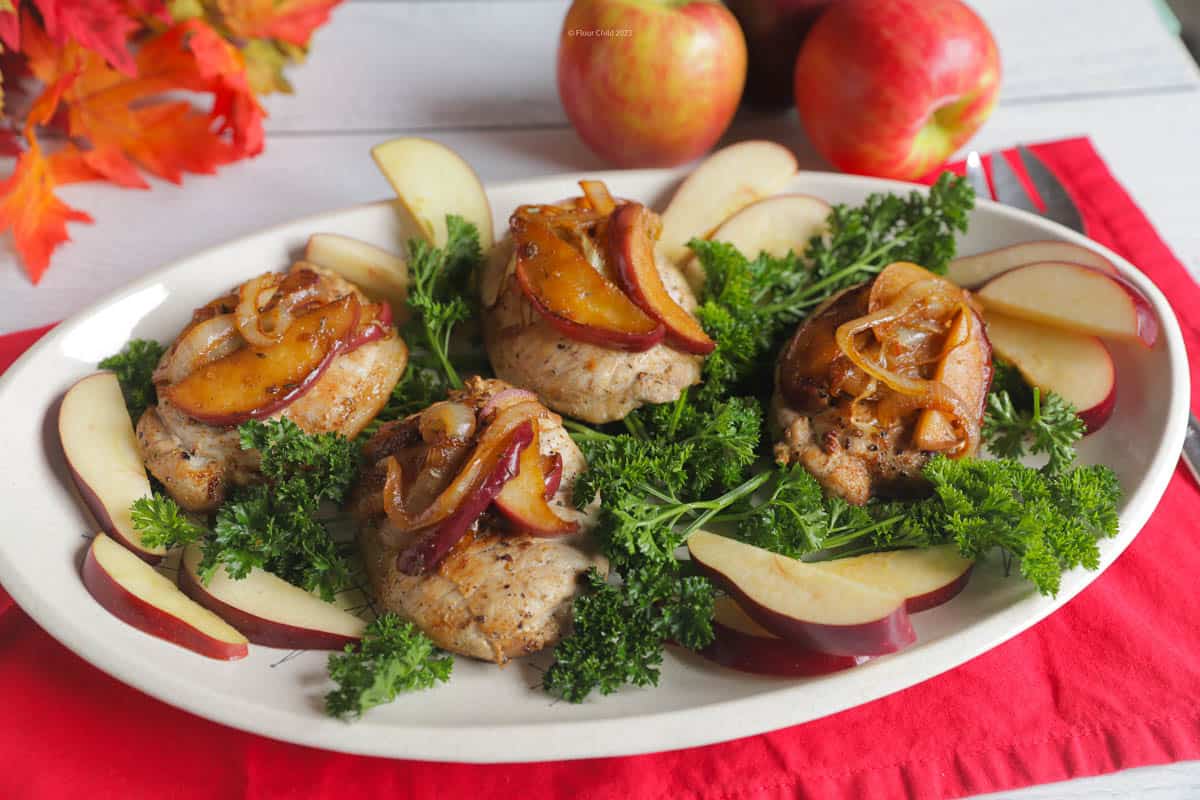 Boneless apple cider pork chops on a serving platter, topped with cooked apples and surrounded by springs of fresh parsley.