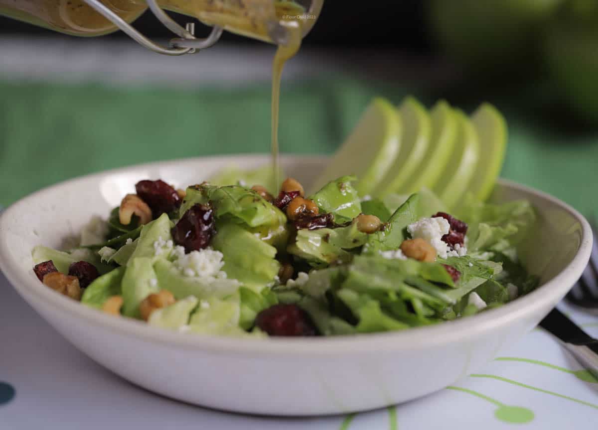 Candy apple salad in a white salad bowl, with apple cider vinaigrette being poured on top from a small glass pitcher.