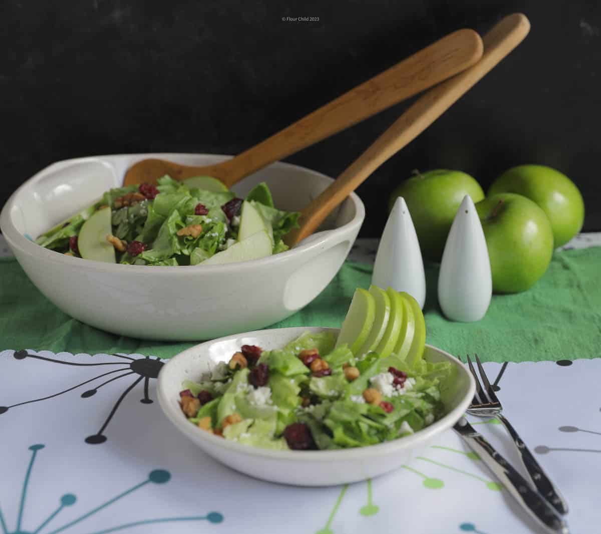 Candy Apple Salad in a white salad bowl, with a serving bowl with wooden serving spoons behind it.