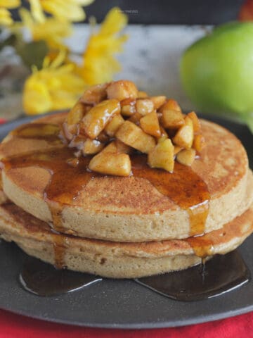 Two large cinnamon apple pancakes on a black plate with apple topping and maple syrup running down the sides.