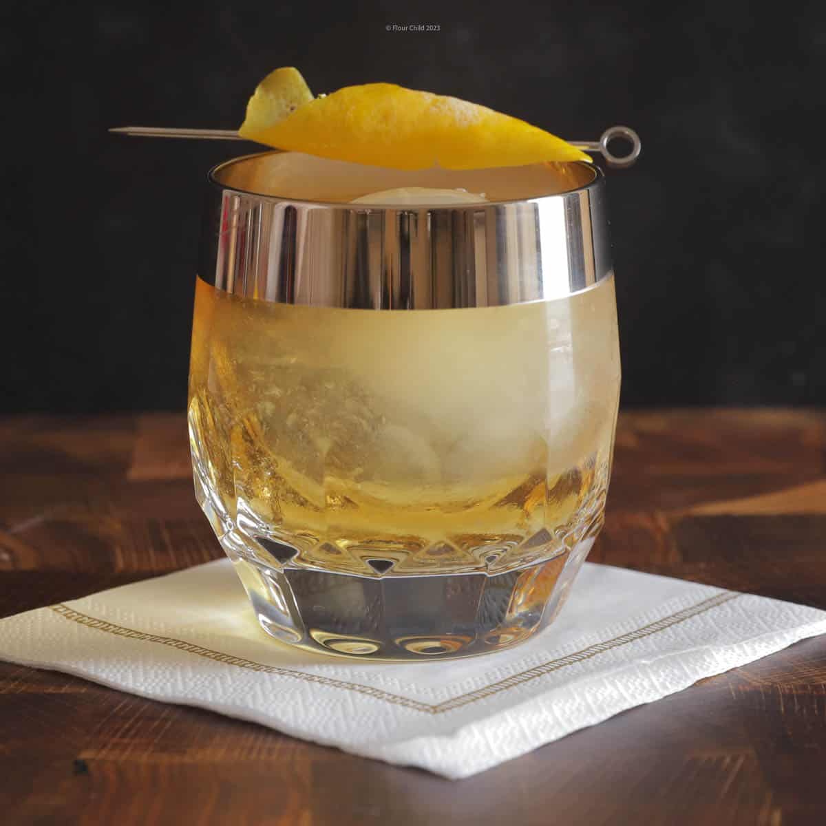 A rusty nail cocktail in a round rocks glass with a slice of lemon on a white bar napkin