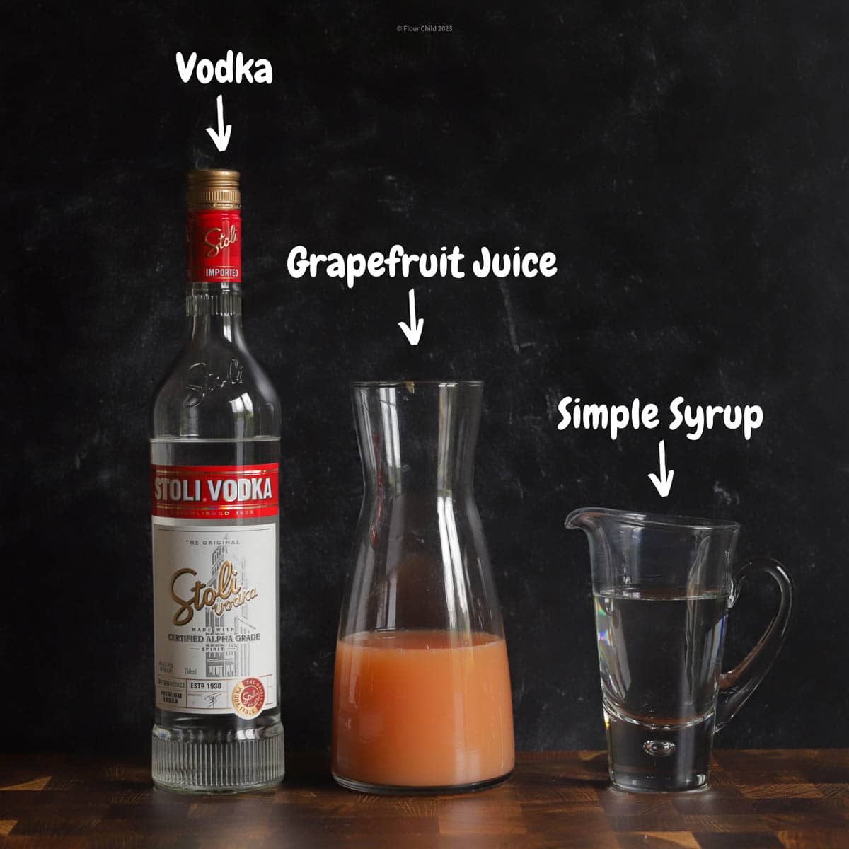 A bottle of vodka and a carafe of grapefruit juice and a glass of simple syrup on a black background
