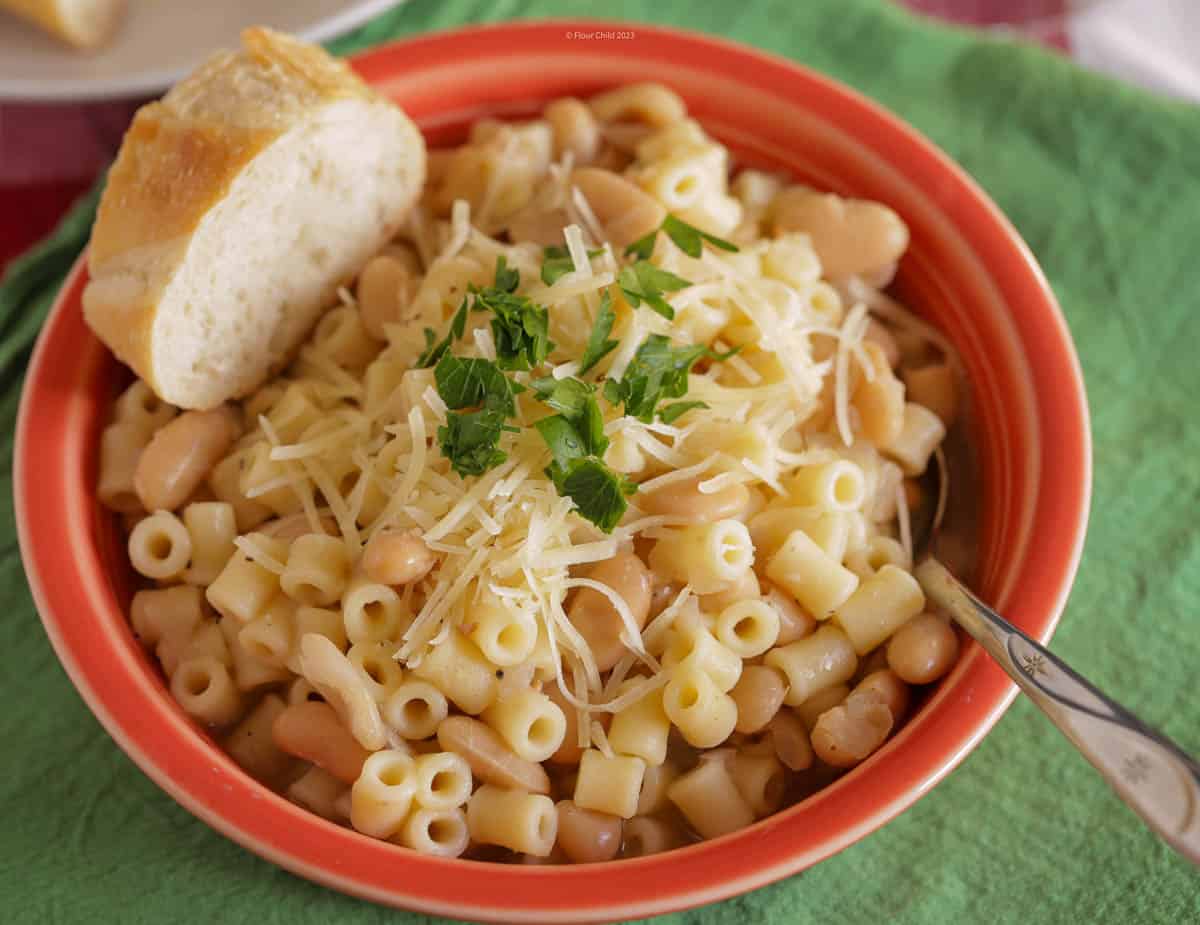 Pasta fagioli in a red bowl with a silver spoon inside, a piece of bread on the rim, and some grated parmesan and chopped Italian parsley on the top.