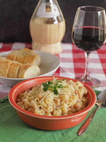 A bowl of pasta fagioli witting on a red checkered tablecloth with a green napkin, a spoon, a bowl with slices of French bread, a glass of red wine, and a bottle of chianti in the background.
