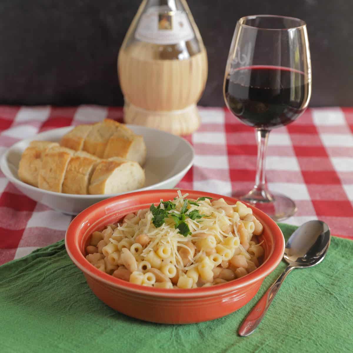 A bowl of pasta fagioli witting on a red checkered tablecloth with a green napkin, a spoon, a bowl with slices of French bread, a glass of red wine, and a bottle of chianti in the background.