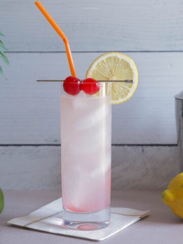 A Rum Collins cocktail on a white background with limes and lemons and two red cherries in a slim glass.