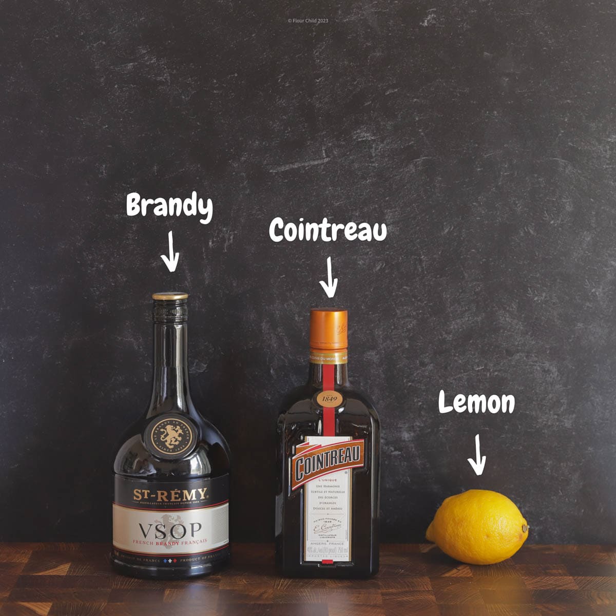 Ingredients for a sidecar cocktail include bottles of brandy, orange liqueur and an orange shown on a black background.