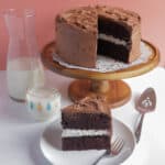 Hershey's Black Magic Dark Chocolate Cake on a cake stand, with a slice cut out and sitting on a white dessert plate next to a glass of milk.